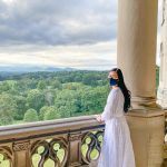 An Asian woman with long hair and a black face mask is wearing a long white dress and looking out on from a balcony at Biltmore Estate in Asheville, North Carolina