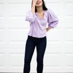 An Asian woman with long hair is laughing while touching her face. She's wearing a lilac peplum top, jeans, and strawberry crocs.