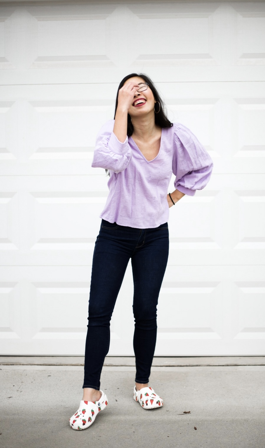 An Asian woman with long hair is laughing while touching her face. She's wearing a lilac peplum top, jeans, and strawberry crocs.