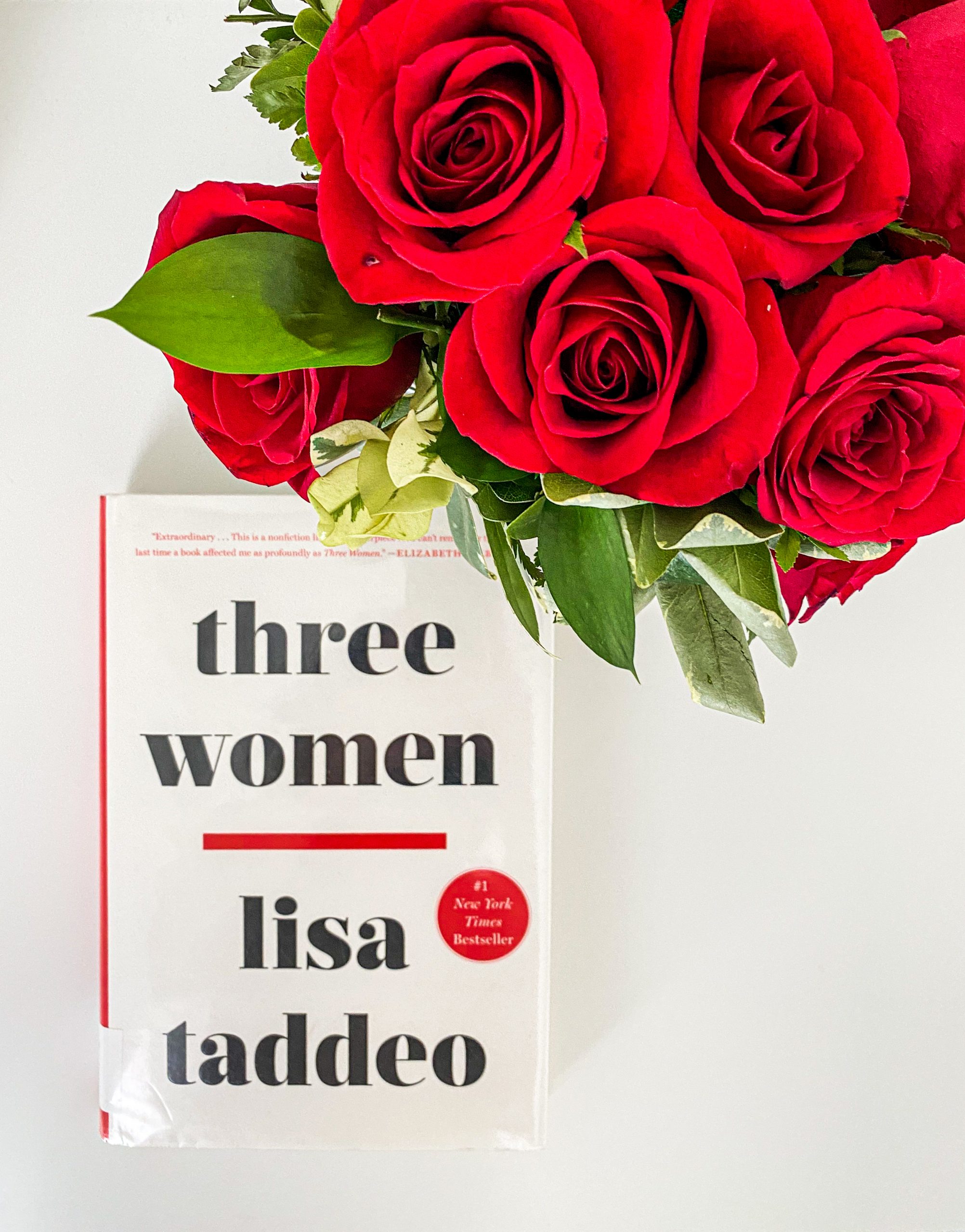 A white book titled Three Women by Lisa Taddeo pictured with a bouquet of red roses