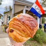 A chocolate croissant being held up in front of a building with a French Flag