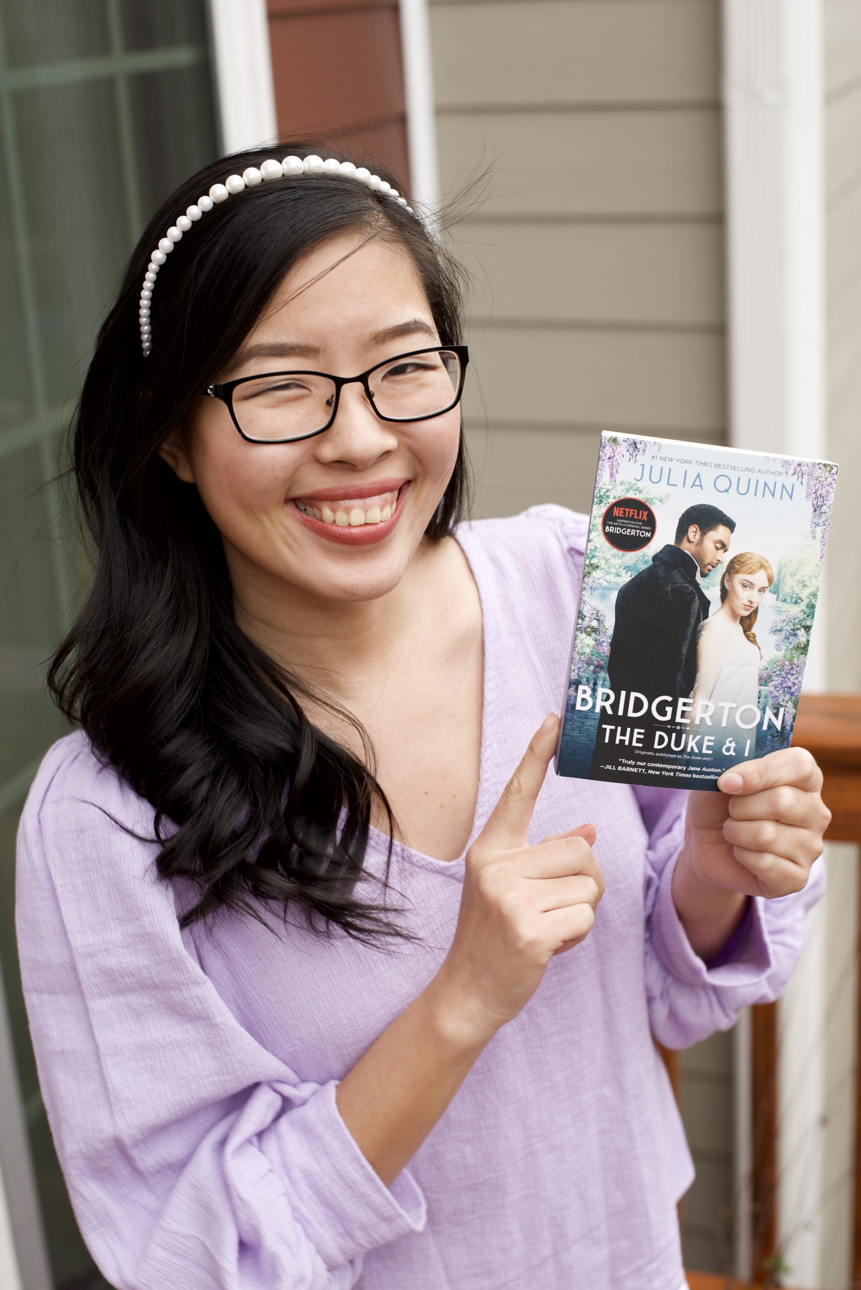An Asian woman wearing a lilac shirt and pearl headband holding the first book of Bridgerton Series "The Duke and I" by Julia Quinn.